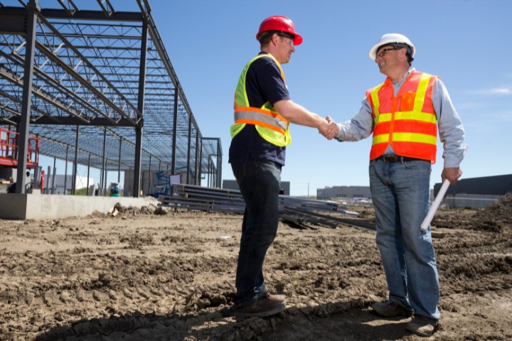 Contractor and project manager in safety gear shaking hands in front of a steel framework under construction