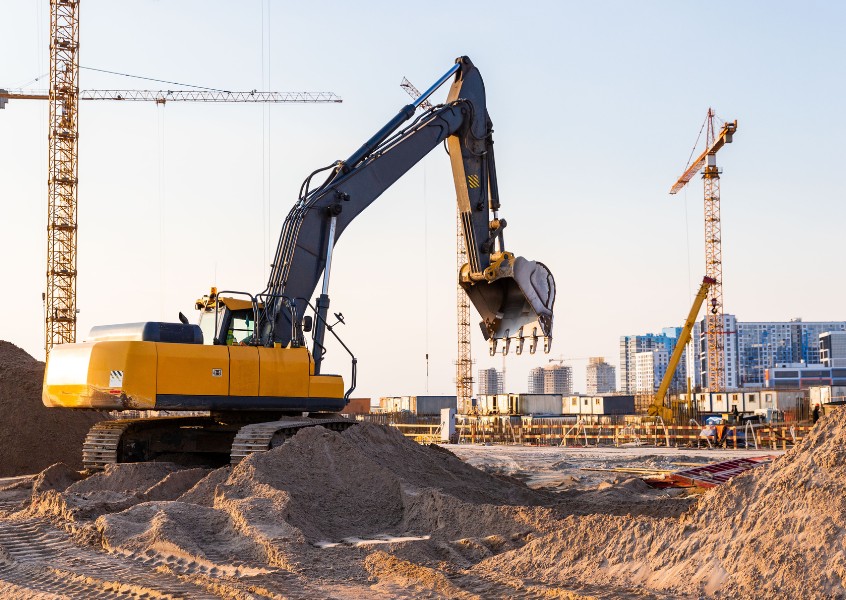 A large yellow excavator moving soil at an urban construction site, surrounded by cranes and high-rise buildings